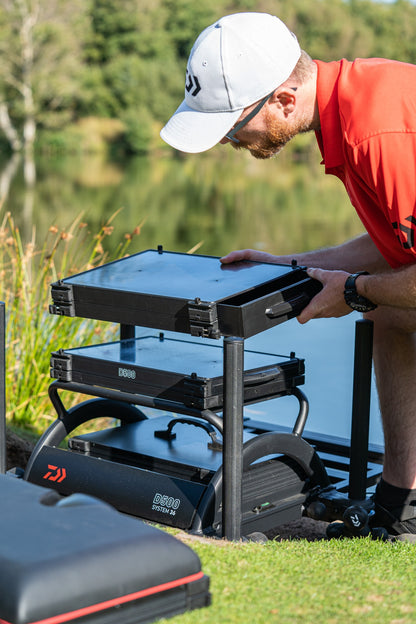 Мъж с червена риза настройва платформата DAIWA D500SB - D36 на риболовно място до вода.