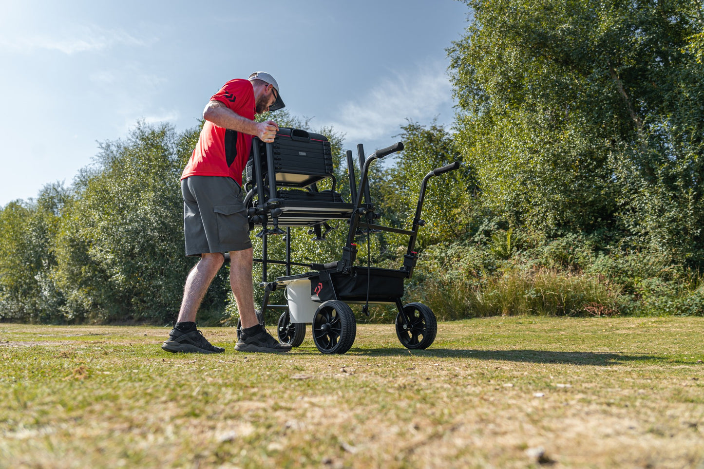 Мъж носи количка за багаж Daiwa DAIWA FOLD N GO MATCH BARROW с рибарски такъми на открито.