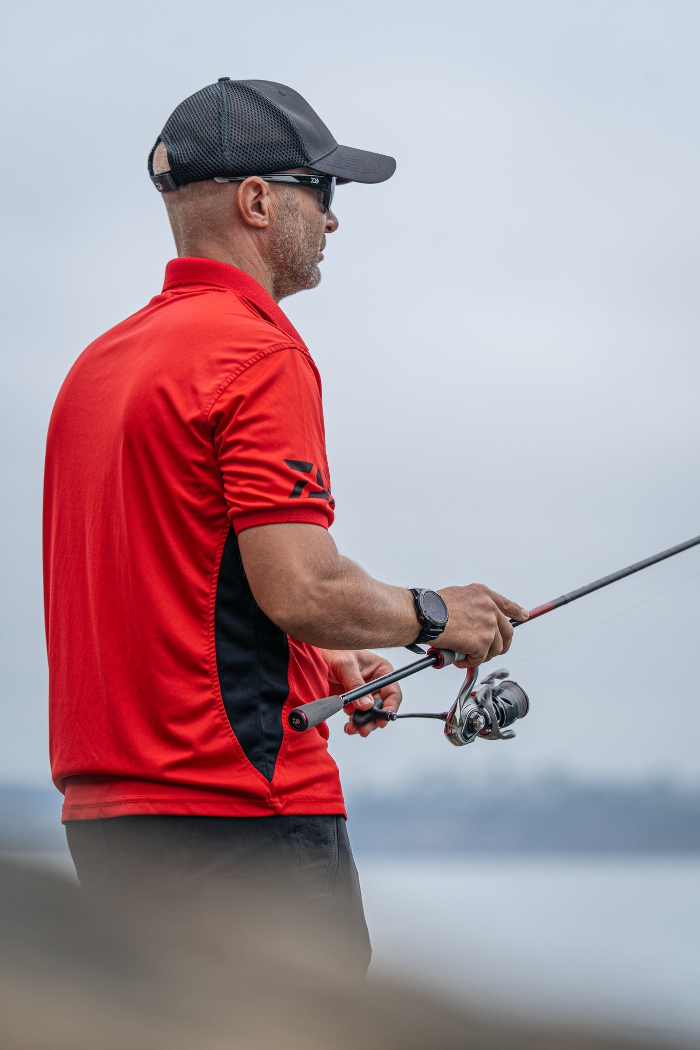Рибар с макара DAIWA 25 CALDIA FC LT и въдица, облечен в червена риза на брега на водоем.