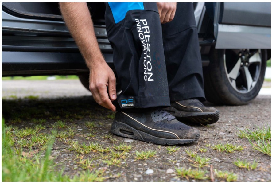 Водоустойчив панталон PRESTON DF ULTRA TROUSERS, удобен за риболов, показан на рибар, обуща и автомобил.