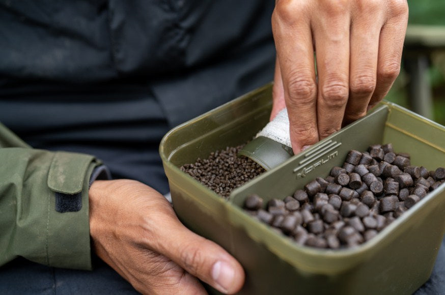Кутия KORUM DIVIDED BAIT TUB с рибарска стръв и затворена преграда в ръката на рибар.