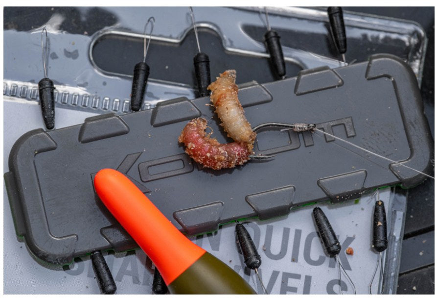 Kуки без контра KORUM XPERT MAGGOT BARBLESS с бял червей и риболовни аксесоари на работната повърхност.